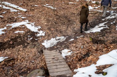 İki yürüyüşçü bir yaya köprüsünü geçer ve kış ormanı patikasına tırmanır.