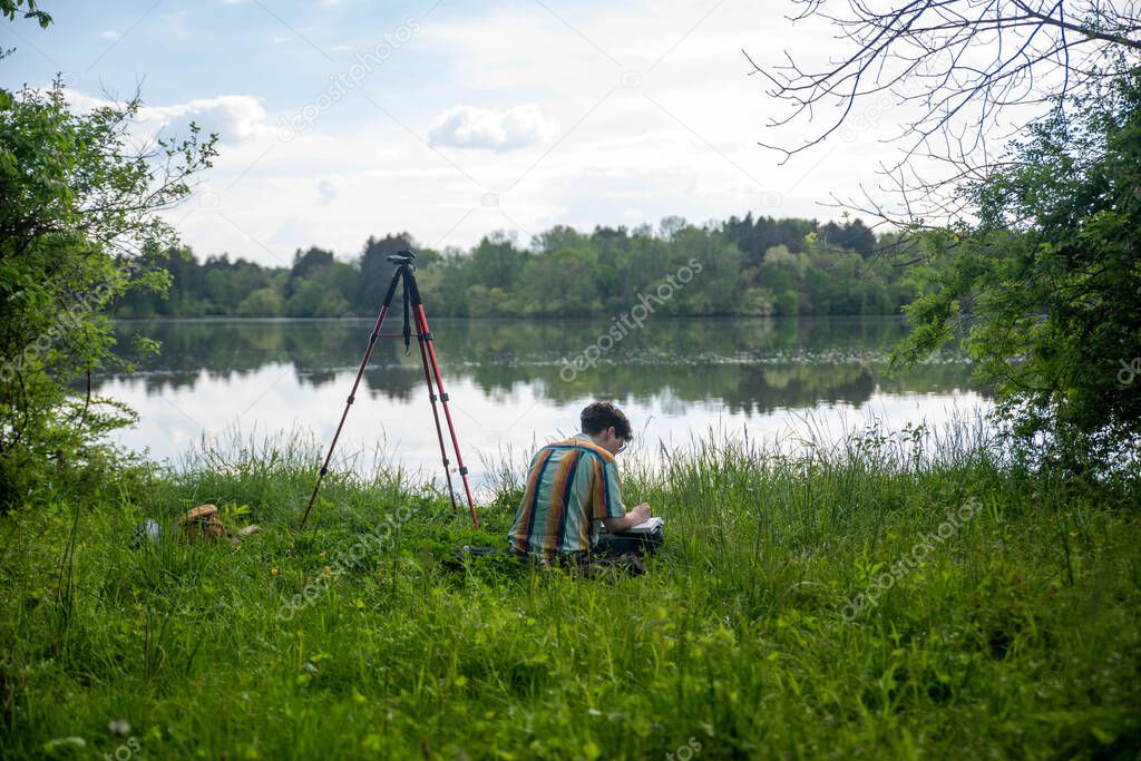 Young man sits in grassy lake shore with photography eqiptment