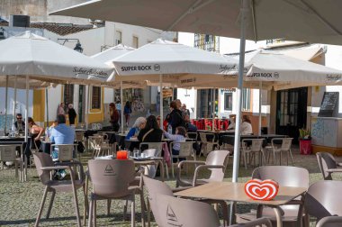 Evora, Portekiz, 11-19-2024 Ortaçağ şehrinde tarihi kaldırım taşları ve dükkanları olan işlek tarihi kaldırım taşları üzerinde gölge şemsiyeli açık hava restoranlarında oturan insanlar..