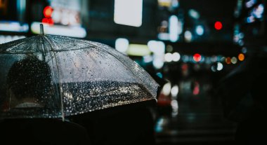 Geceleri şehirde şeffaf bir şemsiyeyle yürüyen bir adam.