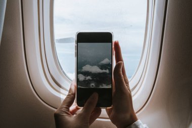 Kadın telefonuyla uçağın camından bulutları yakalıyor.