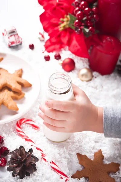 Plätzchen, Zuckerrohr und Milch für den Weihnachtsmann auf dem Tisch vor der Weihnachtsdekoration. Kinderhand nimmt Flasche mit Milch. — Stockfoto