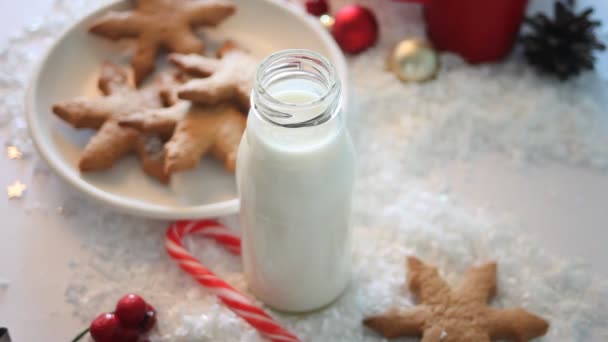 Plätzchen, Zuckerrohr und Milch für den Weihnachtsmann auf dem Tisch vor der Weihnachtsdekoration. Kinderhand nimmt Flasche mit Milch. — Stockvideo