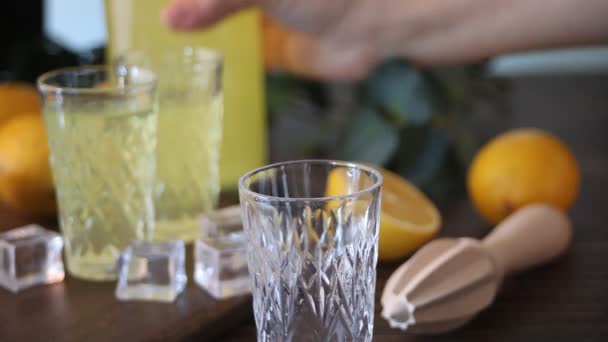 Mão humana derramando limoncelo licor de limão bebida italiana em vidro na mesa de madeira com garrafa e fritas de limão — Vídeo de Stock