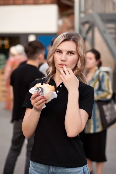 可愛いです若いですブロンド女性食べるハンバーガー屋外で通り — ストック写真