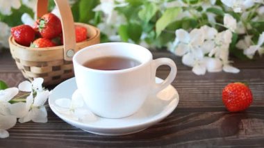 Kadın eli bir fincan çay alır. Yanında çilek ve beyaz elma çiçeği dalıyla birlikte sepet. Bahar konsepti.