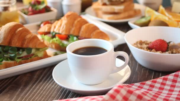 Woman hand pouring milk in to a cup of coffee. Croissant sandwiches with ham and cheese, boiled eggs, muesli and toasts on the table. Breakfast concept. — Vídeo de stock