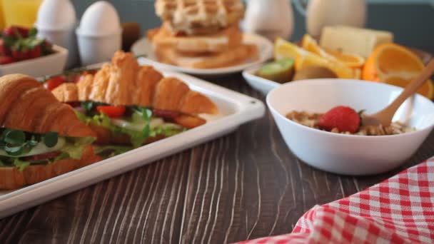 Woman hand put a cup of coffee on the table. Nearby baking sheet with croissant sandwiches with ham, cheese and greens, boiled eggs, muesli and toasts. Breakfast concept — 图库视频影像