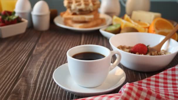 Woman hand put a baking sheet with croissant sandwiches with ham, cheese and greens on the table. Nearby cup of coffee, boiled eggs, muesli and toasts. Breakfast concept. — Vídeos de Stock