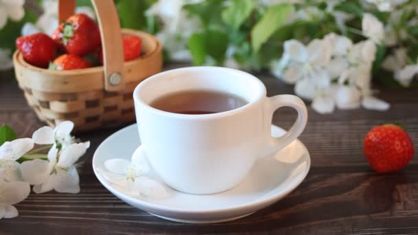 Petals of white apple tree falling in to and nearby cup of black tea. Spring concept. Basket with strawberries on the background. — Stock Video