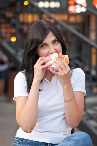 Pretty young brunette woman eating hamburger outdoor on the street — Fotografia de Stock