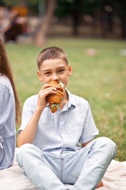 Çocuk yaz tatilinde piknikte oturuyor ve yeni pişmiş kruvasan sandviçi yiyor.