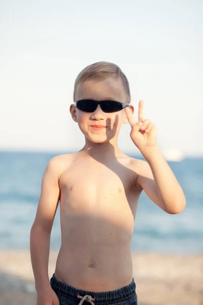 Portrét s chlapečkem v slunečních brýlích stojící na pláži s rozmazaným pozadím u moře — Stock fotografie