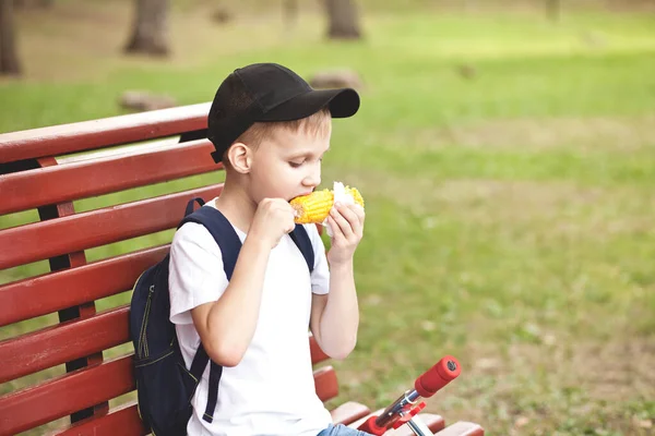 Menino sentado em um banco ao ar livre em um parque e comer milho cozido fresco. — Fotografia de Stock