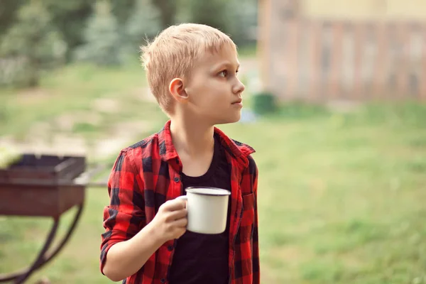 Miúdo adolescente de camisa quadriculada a segurar uma chávena de bebida. Viajar, caminhar, férias, conceito de acampamento. — Fotografia de Stock