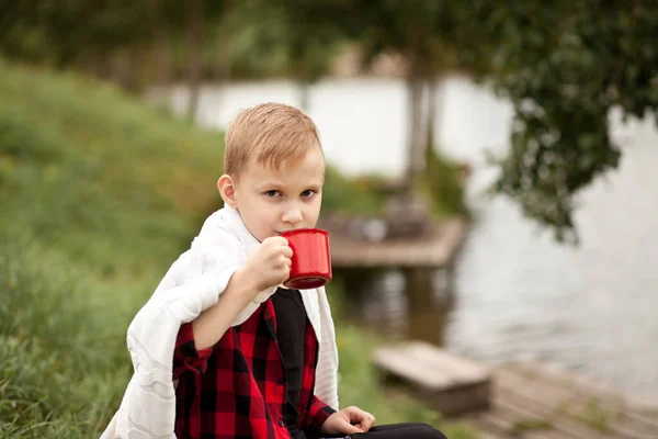 Teen chlapec kluk v kostkované košili drží šálek nápoje v blízkosti rybníka nebo jezera. Cestování, túra, dovolená, koncept kempování. — Stock fotografie