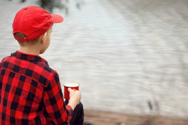 Niño adolescente con camisa a cuadros sosteniendo una taza de bebida cerca de un estanque o lago. Viaje, caminata, vacaciones, concepto de camping. —  Fotos de Stock
