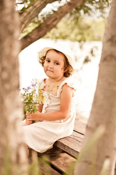 Piccola ragazza felice seduta con un mazzo di fiori selvatici. Estate vita di campagna. — Foto Stock