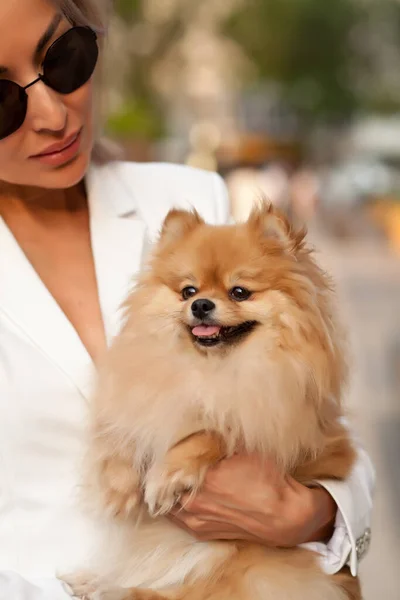 Hermosa mujer rubia en gafas de sol y traje blanco abrazando pequeño perro mascota en un parque al aire libre —  Fotos de Stock