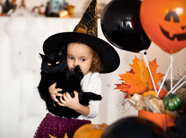 Śmieszne dziecko dziewczyna w strój czarownicy na Halloween z czarnym kotem w rękach. — Zdjęcie stockowe