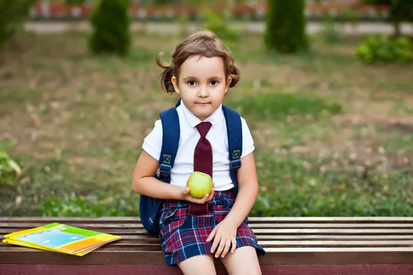 Kleines süßes Schulmädchen in Uniform sitzt auf einer Bank und isst einen Apfel — Stockfoto