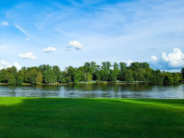 Manzara nehri bir kıyısı yeşil çimlerle kaplıdır diğer ağaçlarda güzel mavi gökyüzü ve birkaç beyaz bulut büyür.