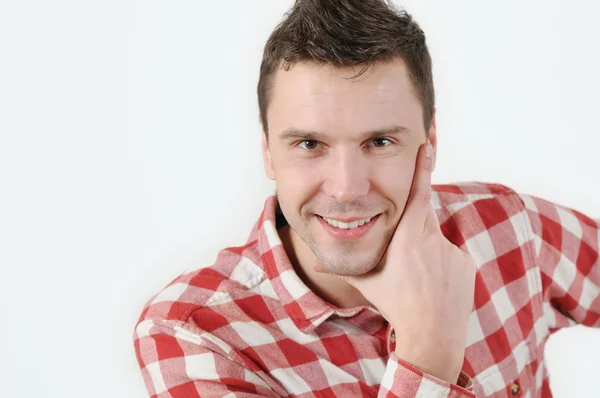 Smiling young man in hipster shirt holding hand on chin and standing against white background — Stock Photo, Image