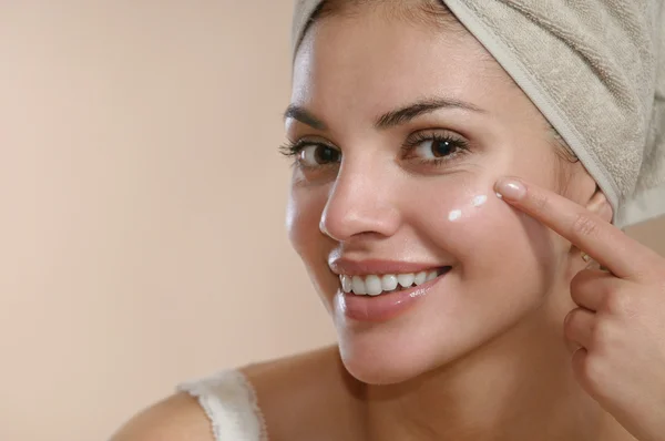 Belle jeune femme enveloppée dans une serviette étalant de la crème sur son visage — Photo