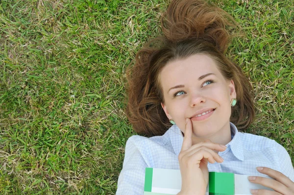 Cute thinking girl with book lying on the grass in the park — Stock Photo, Image
