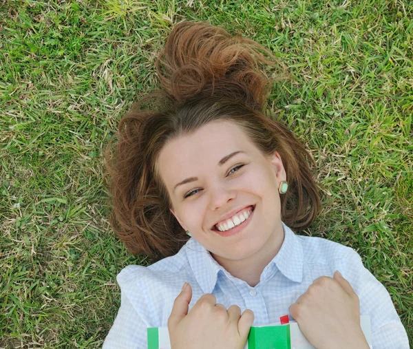 Glada leende flicka med boken liggande på gräset i parken — Stockfoto