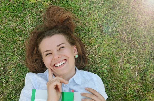 Happy brett leende flicka med boken liggande på gräset i parken — Stockfoto