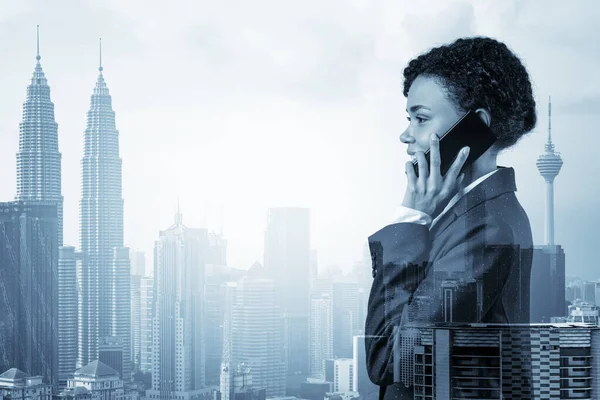 Успішно посміхаючись чорній афроамериканській діловій жінці в костюмі енергійної розмови по телефону, Куала-Лумпур цитайскейп. Концепція консультантів як розв'язувачів проблем. KL skyscrapers Подвійний вплив. — стокове фото
