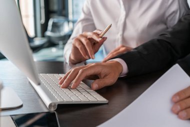 Ofis müdürü klavyeye yazı yazıyor, klavyeyle ellerin yakın plan görüntüsü. Beyaz takım elbiseli kadın yönetici elinde kalemle bilgisayarlı bir adamın yanında oturuyor, yüzü yok. Takım çalışması kavramı