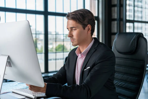 Der junge Geschäftsmann trägt schwarzen Anzug und rosa Hemd und sitzt mit dem Computer am Tisch. Büroleiter arbeitet in modernem Bürogebäude mit Glasfenstern verschwommenem Hintergrund, Arbeitskonzept — Stockfoto