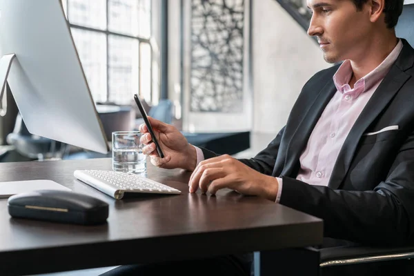 Der junge Geschäftsmann schaut auf das Telefon, trägt einen schwarzen Anzug und sitzt mit dem Computer am Tisch. Büroleiter mit Telefon arbeitet in modernem Büro mit Glasfenstern verschwommenem Hintergrund, Arbeitskonzept — Stockfoto