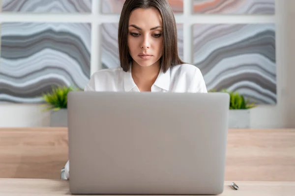 Office manager woman wearing white shirt, sitting to table with laptop, front view. Concentrated office worker looking at the screen in big light office room. Concept of work