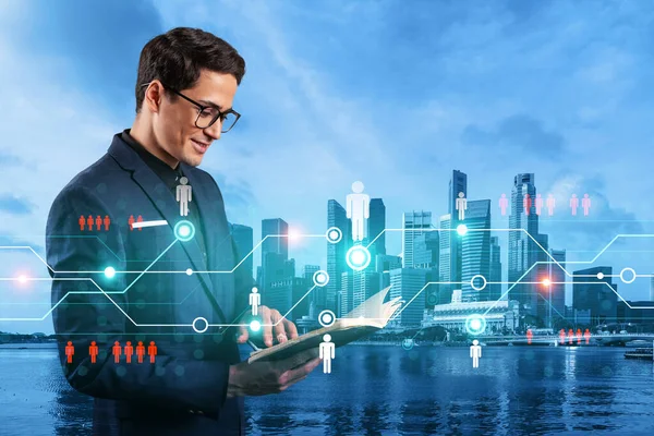 Handsome eastern HR director at international company is looking into personal organizer to plan business meeting to recruit highly qualified specialists. Social media hologram icons over Singapore.