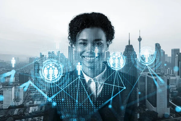 Smiling black woman HR director at international company is thinking about recruitment of highly qualified specialists. Women in business concept. Social media hologram icons over Kuala Lumpur.