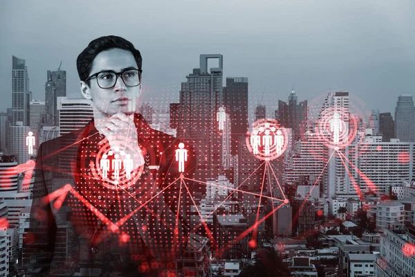 Handsome eastern HR director at international company is thinking about efficient strategy to recruit highly qualified specialists. Social media and marketing hologram icons over Bangkok background.