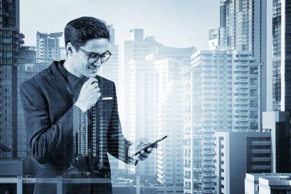 Joven hombre de negocios guapo en traje y gafas usando el teléfono y pensando en cómo abordar el problema, nuevas oportunidades de carrera, MBA. Bangkok en el fondo. Doble exposición. — Foto de Stock
