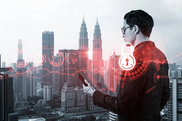 Un joven y apuesto desarrollador de ciberseguridad oriental explora nuevos enfoques para proteger la información confidencial de los clientes mediante el teléfono. Iconos de bloqueo de TI sobre fondo de la ciudad de Kuala Lumpur. — Foto de Stock