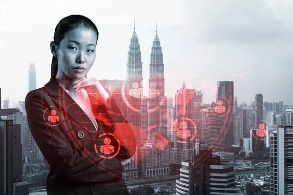 Attractive young Asian HR director at international company is thinking about recruitment of highly qualified specialists. Women in business concept. Social media hologram icons over Kuala Lumpur.