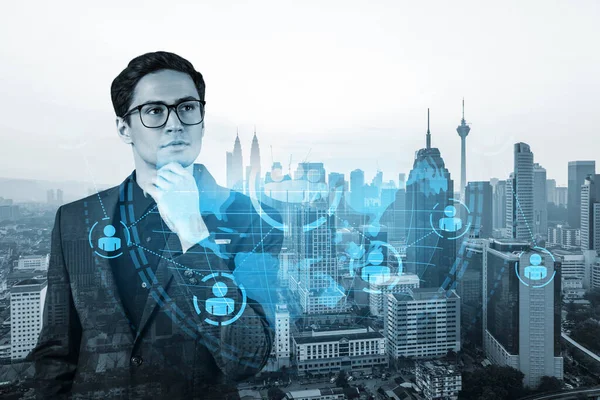 Handsome eastern HR director at international company thinking about efficient strategy to recruit highly qualified specialists. Social media and marketing hologram icons over Kuala Lumpur background