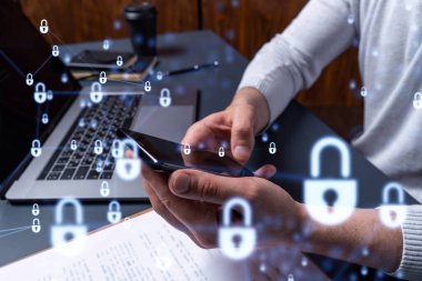 Bir programcı, bir siber güvenliği hacker saldırılarından korumak ve müşterilerinin gizli verilerini kaydetmek için akıllı telefonuyla internete giriyor. Yazan ellerin üzerine asma kilit hologramı simgeleri.