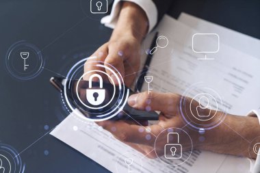 Resmi görevli bir işadamı siber güvenlikte risk olasılığını önlemek ve telefonda detayları kontrol etmek için sözleşmeyi okuyor. Çalışma masasının üzerine asma kilit hologramı simgeleri.