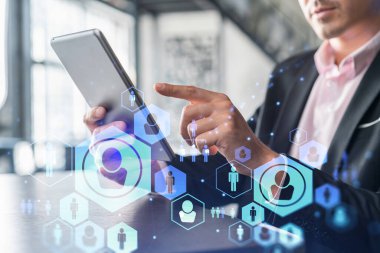 Takım elbiseli bir işadamı uluslararası iş danışmanlığı için yeni adayları kontrol etmek için tablet kullanıyor. HR ve sosyal medya simgeleri modern panoramik ofis geçmişi üzerine.