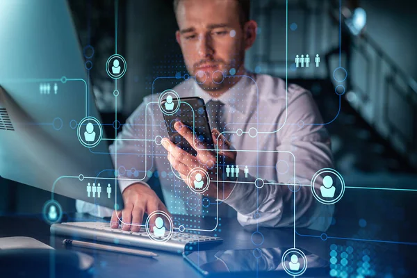 国際的なビジネスコンサルティングのための新しい候補を確認するためにスマートフォンを使用してカジュアルウェアのビジネスマン。HR 、現代のオフィスの背景にソーシャルメディアのアイコン夜の時間. — ストック写真