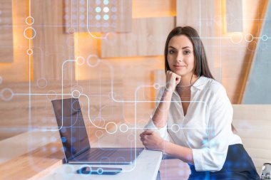 İş yerindeki beyaz tişörtlü çekici iş kadını iş hayatında yeni teknolojiler ima ederek gelişimi optimize etmek için dizüstü bilgisayarla çalışıyor. Ofis geçmişinde yüksek teknoloji hologramı