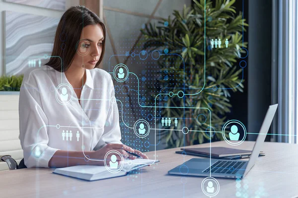 Attractive businesswoman in white shirt at workplace working with laptop to hire new employees for international business consulting. HR, social media hologram icons over office background — Stock Photo, Image