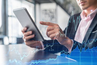 Takım elbiseli bir işadamı, kurumsal finans fonundaki ticaret stratejisini optimize etmek için tablet cihazını kullanıyor. Modern panoramik ofis arkaplanı üzerinde eski grafik hologramı